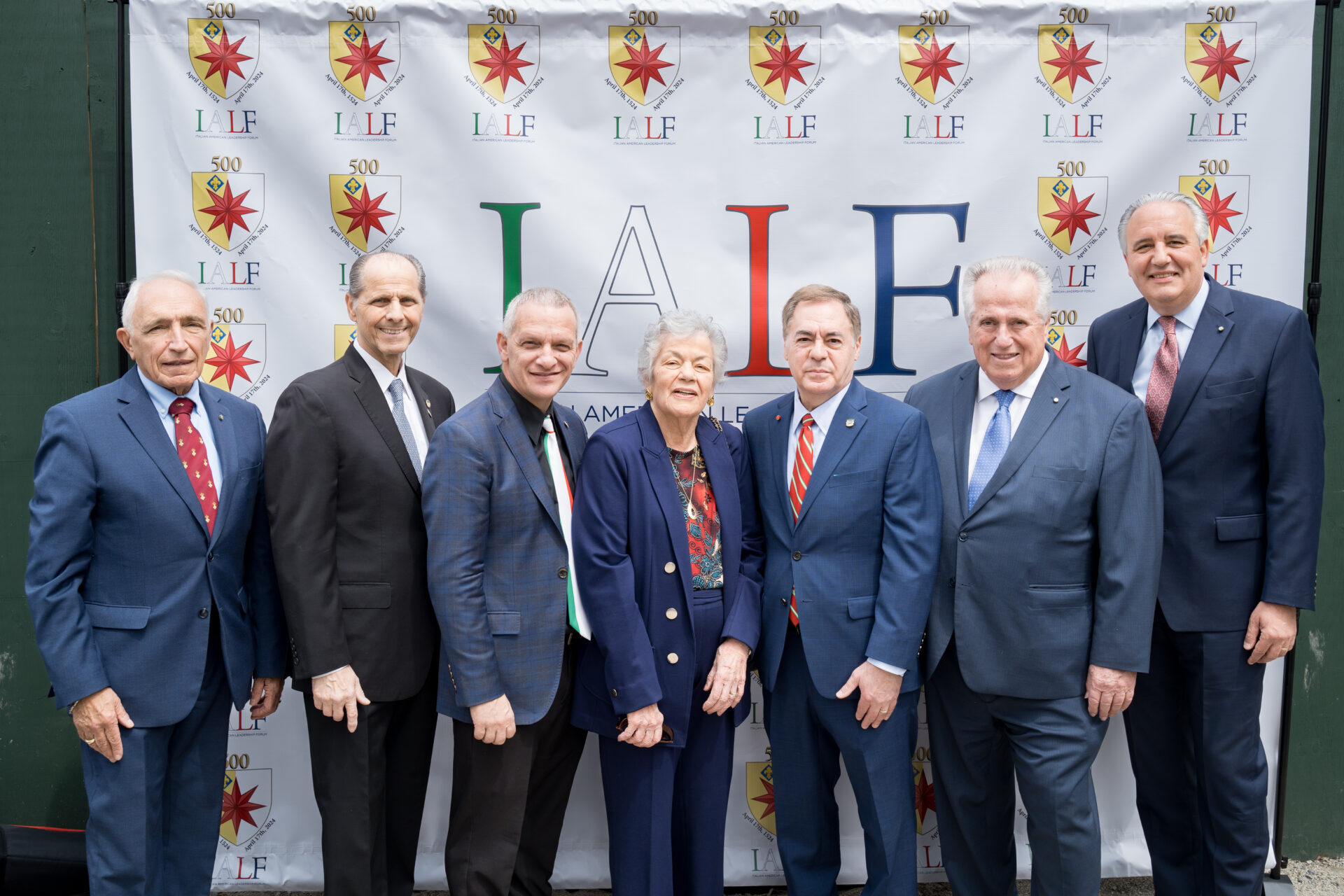 Seven directors from the seven founding organizations of the Italian-American Leadership Forum pose in front of a backdrop that features the organization logos.