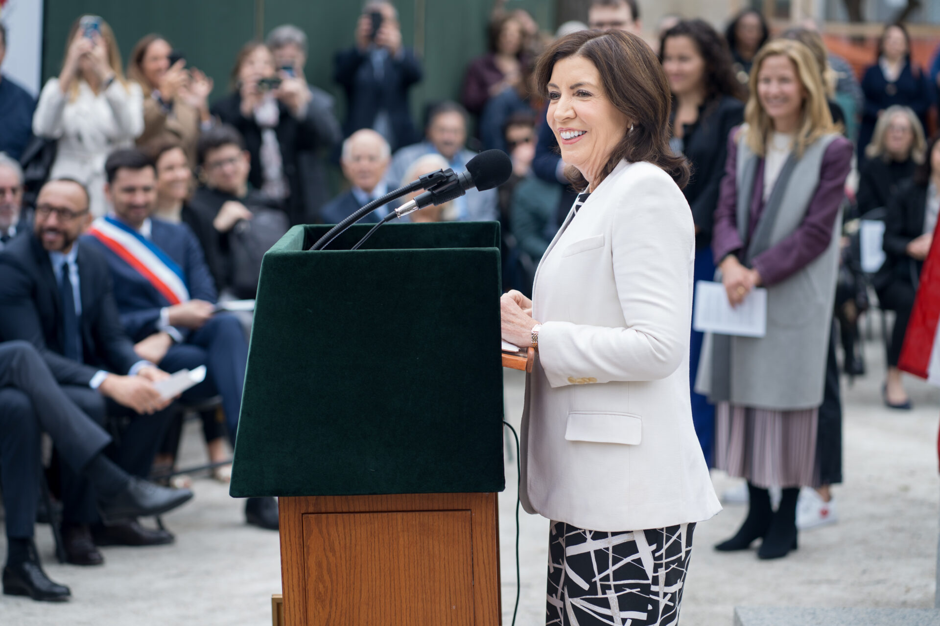 A woman in a white blazer speaks at an outdoor podium with microphones, smiling as she addresses an audience. People in the crowd, some seated and others standing, listen attentively and take photos.