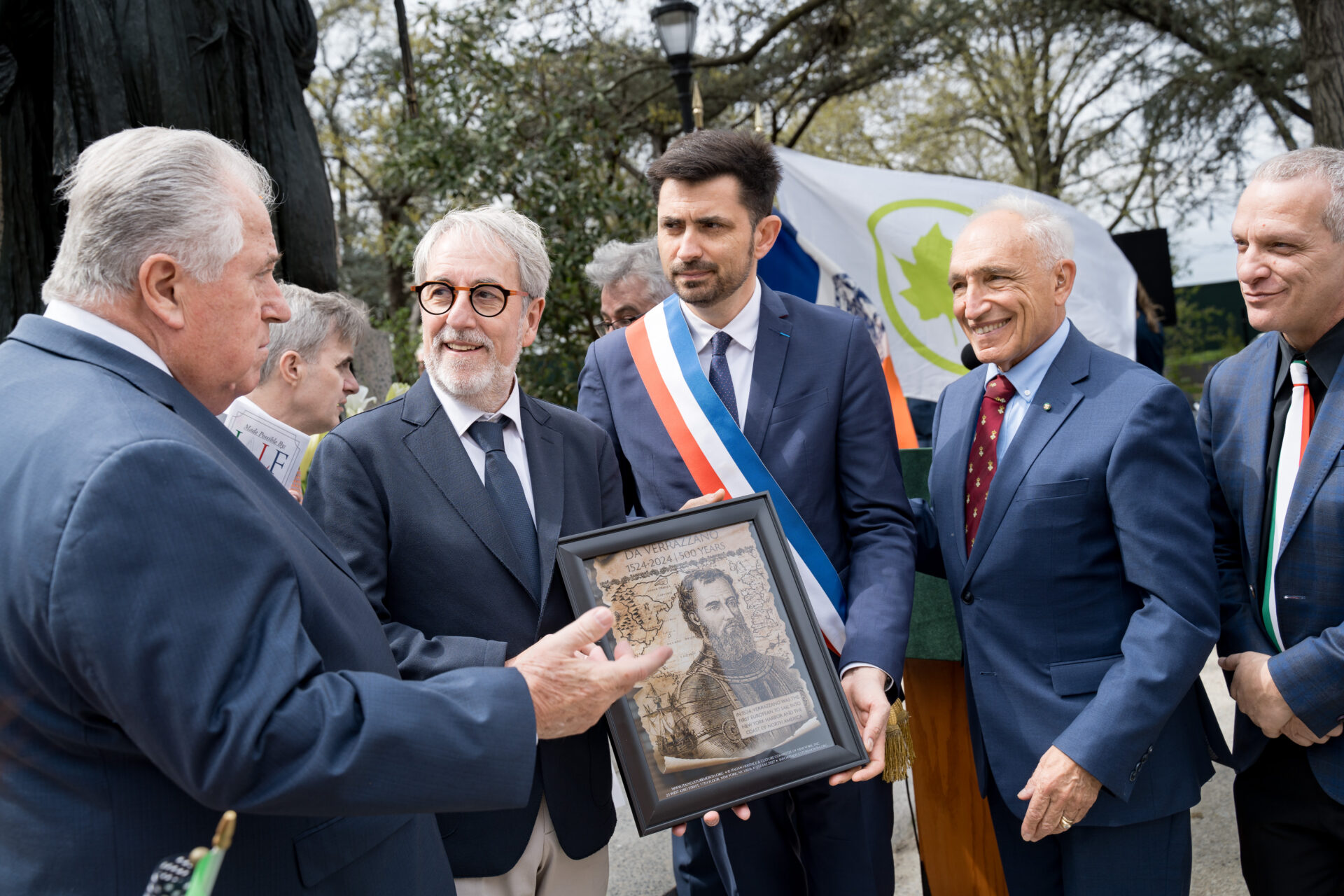 A group of formally dressed men stand outdoors during a ceremony, with one man presenting a framed portrait of explorer Giovanni da Verrazzano to another.