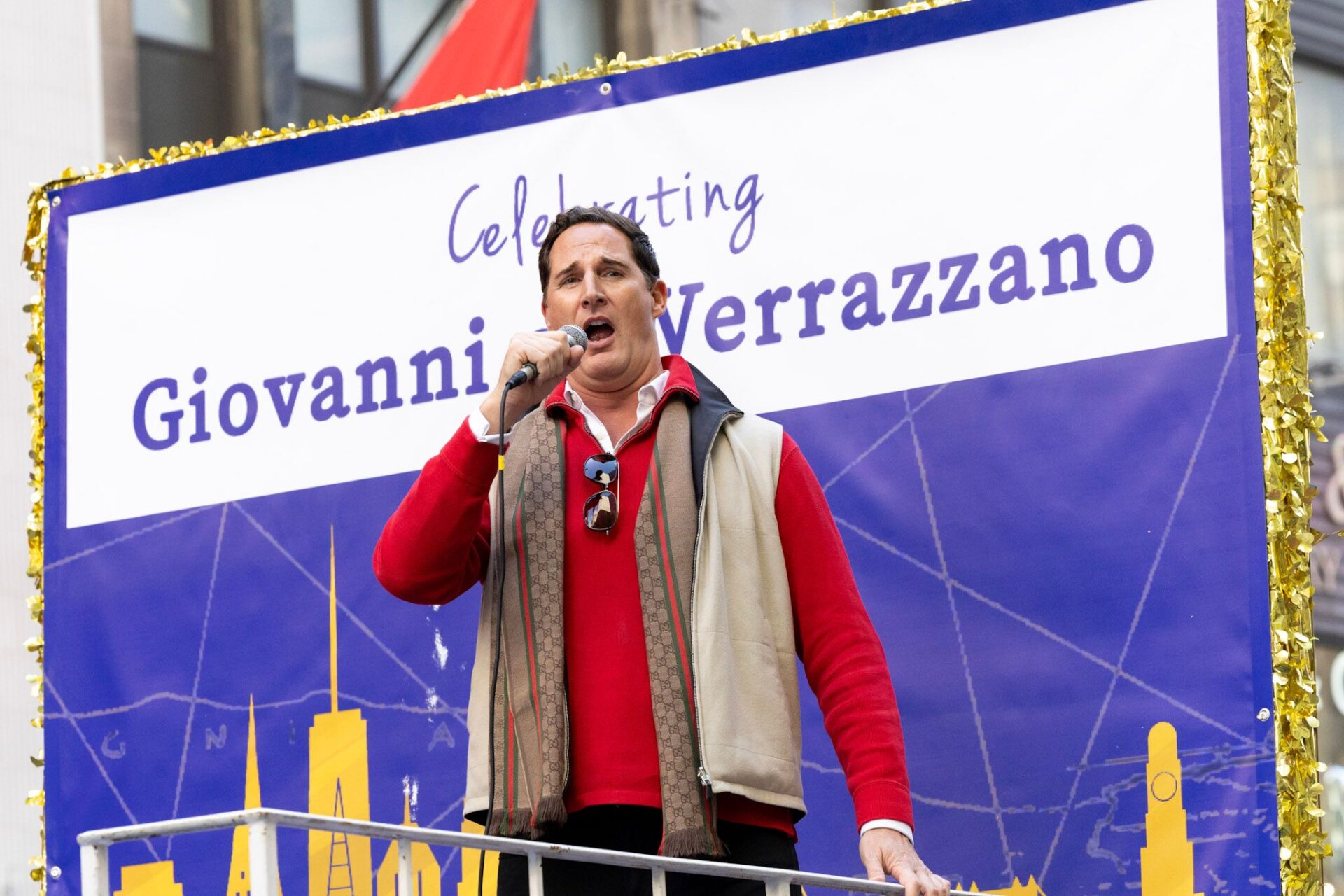 A man wearing a red sweater, beige vest, and scarf speaks into a microphone on stage during a celebration of Giovanni da Verrazzano. A large banner behind him reads “Celebrating Giovanni Verrazzano” with a blue background and gold city skyline design.